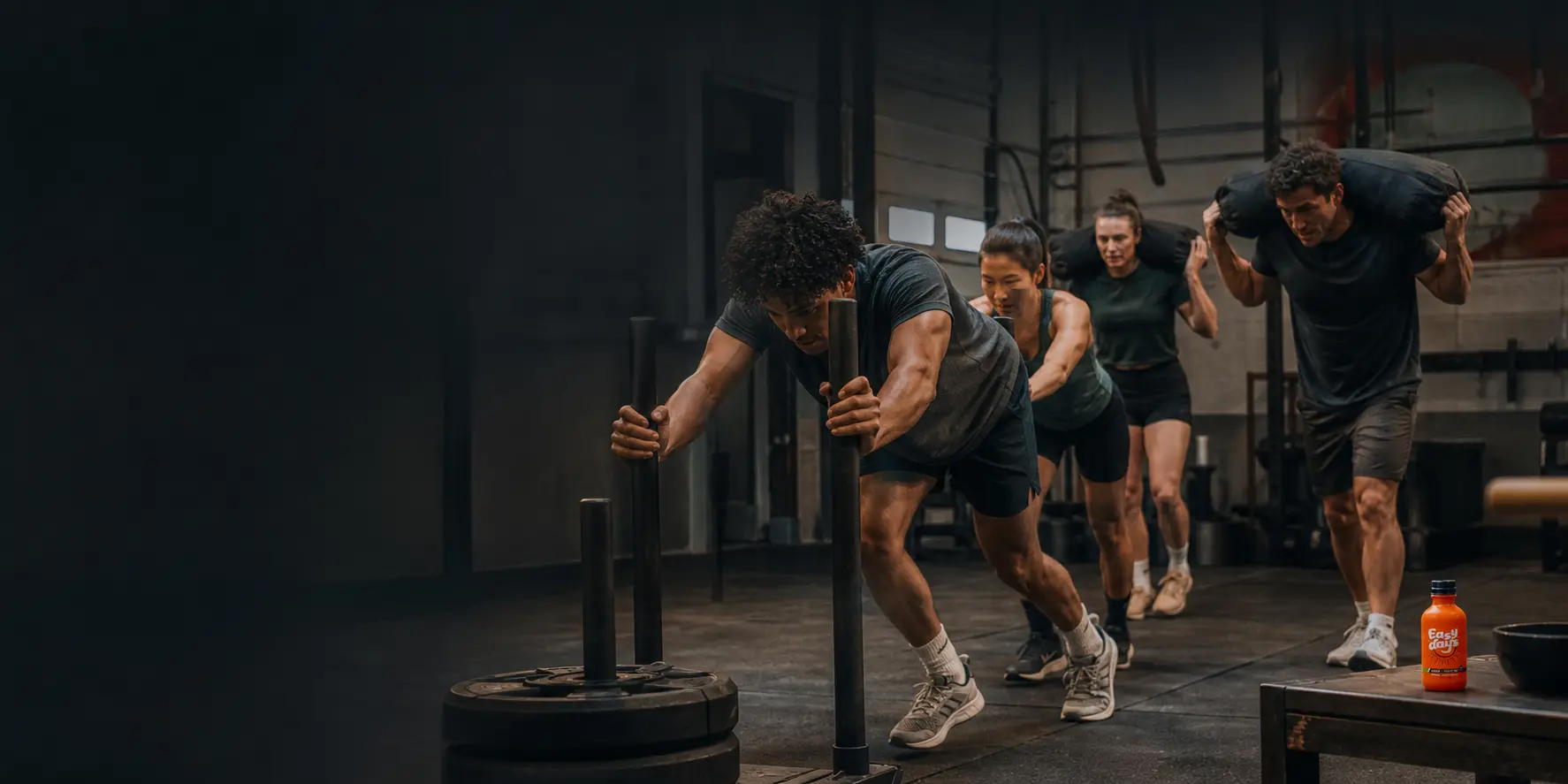 Athletes pushing a sled in a gym with an Easy Days bottle on a bench